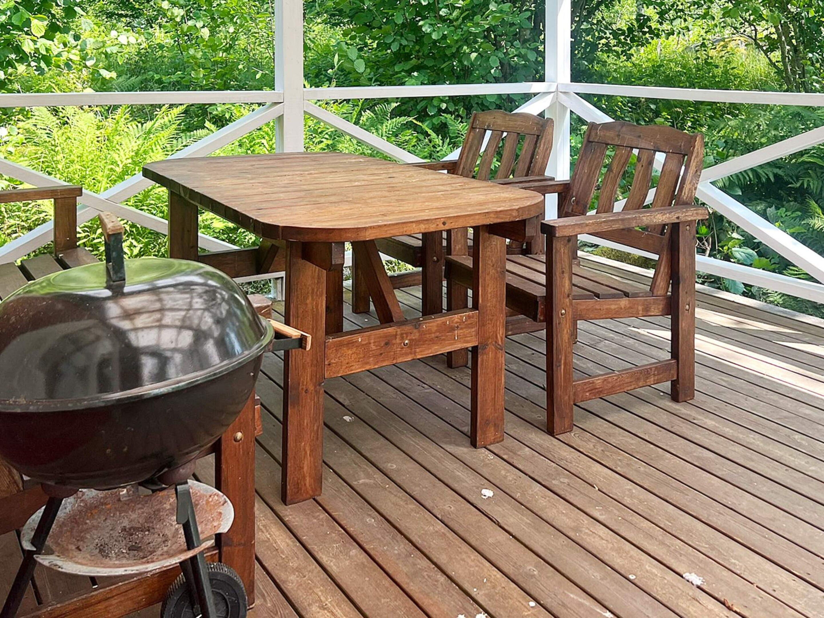 A wooden deck with a table and chairs, a grill, and lush greenery in the background.