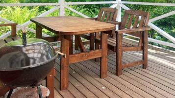 A wooden deck with a table and chairs, a grill, and lush greenery in the background.