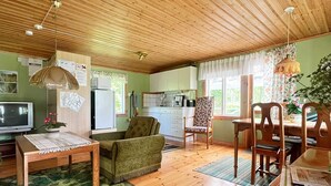 Ein gemütliches Wohnzimmer mit Holzbalkendecke, einem Fernseher, einem Sofa, einem Esstisch mit Stühlen und einer Küche im Hintergrund.