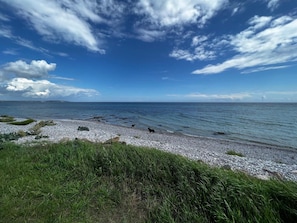 Une plage de galets, avec de l’herbe au premier plan et un ciel dégagé.