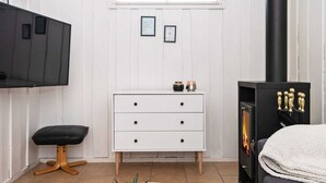 Une chambre moderne avec une commode blanche, un téléviseur noir fixé au mur, un poêle à bois noir, un banc noir et une table ronde blanche agrémentée d’une plante et d’un vase en verre.