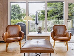 Een kamer met twee leren fauteuils en een bank, een tafel met een lamp, en uitzicht op een tuin door grote ramen.