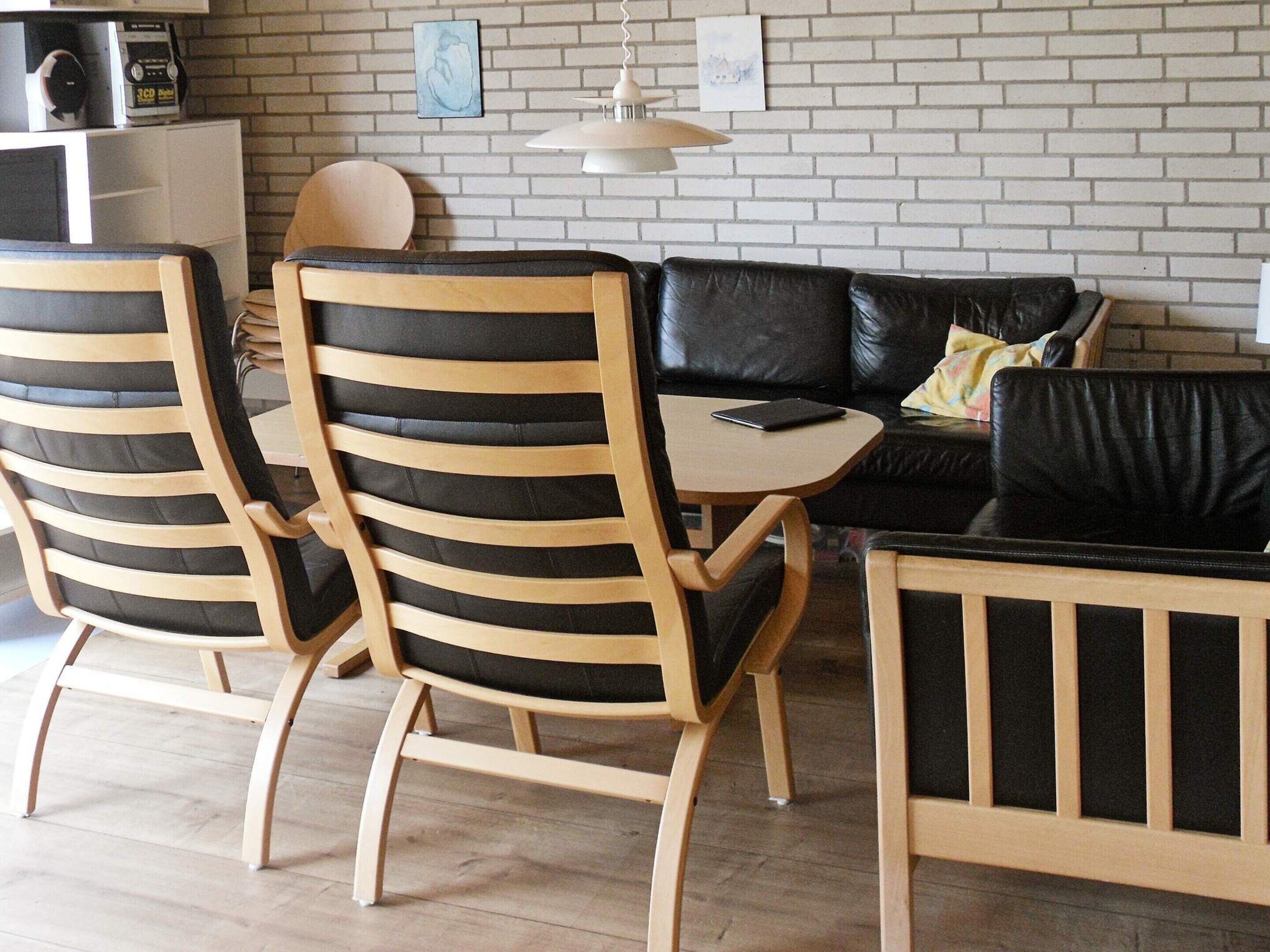 A modern living space with a black leather sofa, wooden chairs with black cushions, a small wooden table, and a white brick wall.