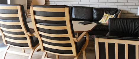 Ein moderner Wohnraum mit einem schwarzen Ledersofa, Holzstühlen mit schwarzen Kissen, einem kleinen Holztisch und einer Wand aus weißen Ziegeln.