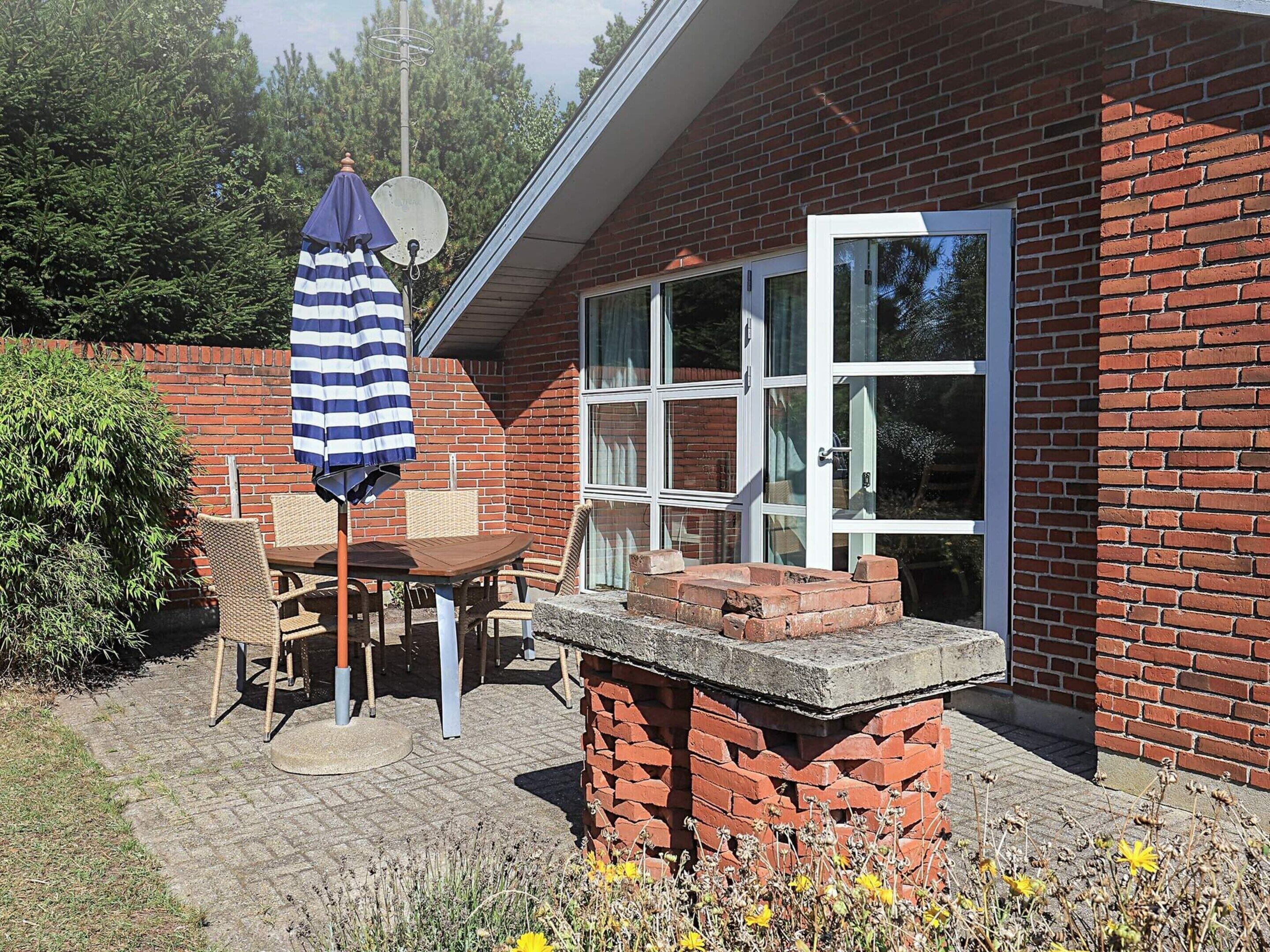 Une maison en briques avec un patio, une table entourée de chaises, un parasol à rayures et une antenne parabolique.
