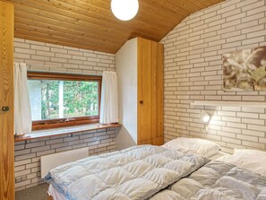A bedroom with a brick wall, wooden ceiling, a window with curtains, a bed with white bedding, and a wall-mounted light fixture. - 4 Person Holiday Home in Vaeggerlose (Vaeggerlose)