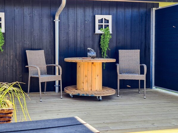 Una terraza de madera con una mesa redonda y dos sillas de mimbre, un muro negro con un ventanal, y una planta colgando de la ventana.