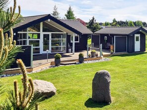 Ein modernes, zweistöckiges Haus mit großen Fenstern, einer Holzterrasse und einem Kiesweg, der zum Eingang führt.