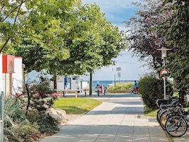 Un sendero que lleva a una playa con un banco y un poste señalizador, bicicletas estacionadas a un lado, y un edificio con fachada roja y blanca.