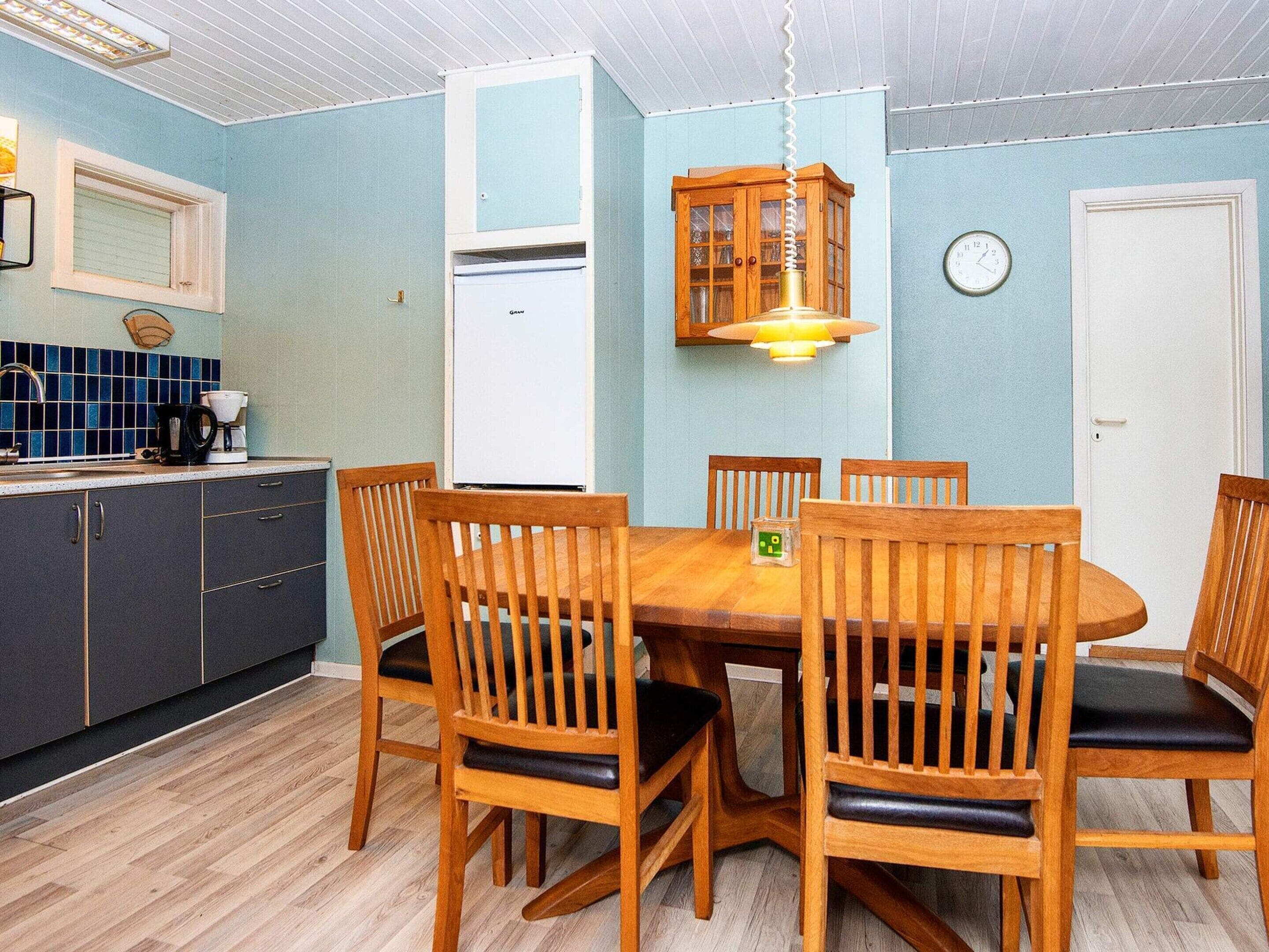 A dining area with a wooden table and chairs, a refrigerator, a clock, and a window with blinds.
