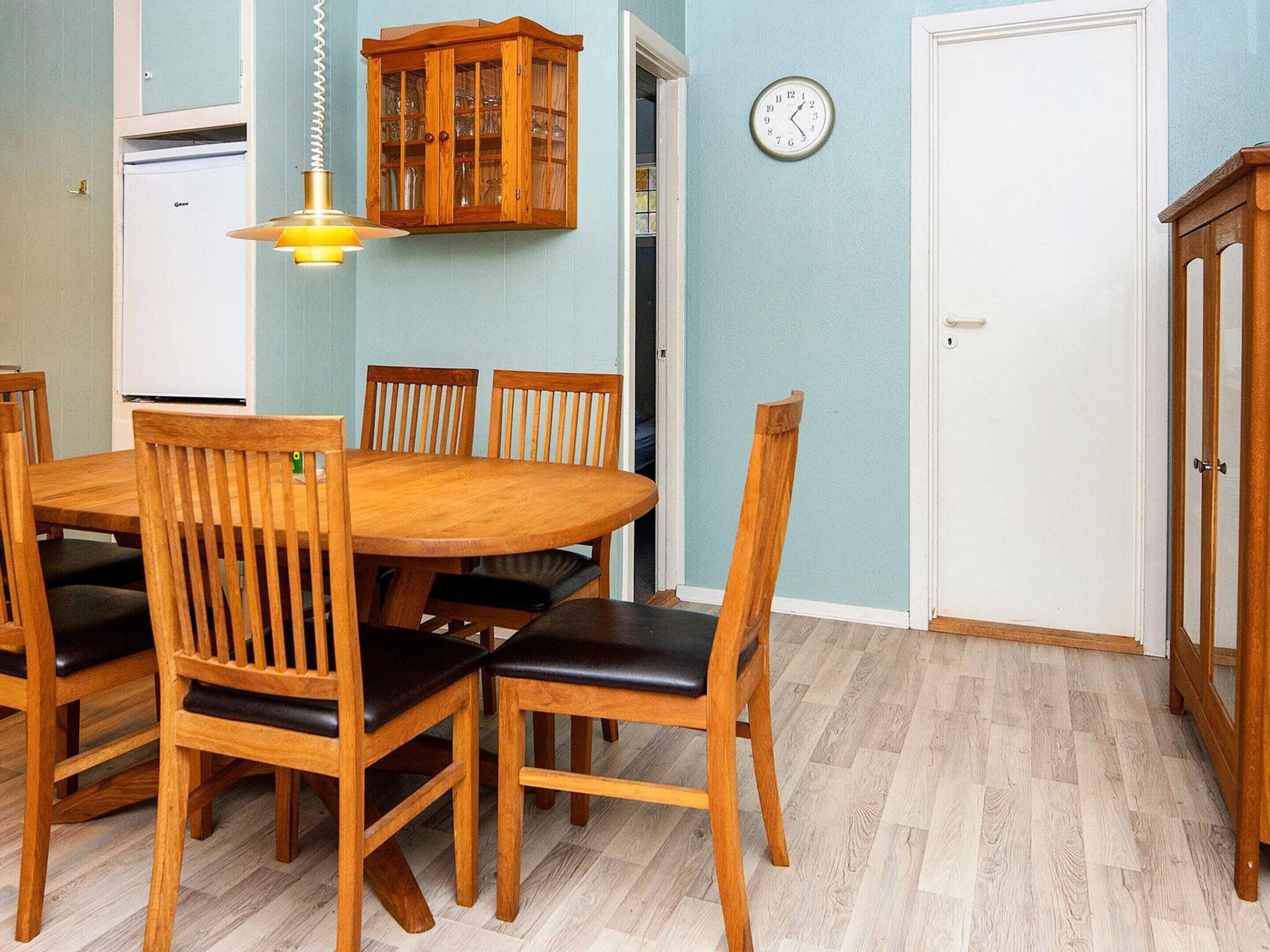 Un comedor con una mesa y sillas de madera, una lámpara colgante, un reloj en la pared y un armario con puertas de vidrio.