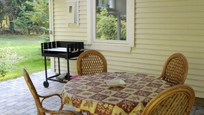 Eine überdachte Terrasse mit Tisch und Stühlen, ein Fenster mit Blick auf Grünflächen und ein kleiner Wagen.