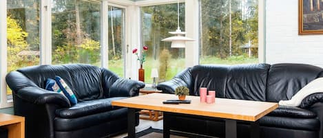 A room with a black leather sofa set, a wooden table, and large windows offering a view of trees.