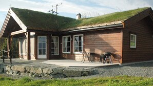 En træhytte med græstag, stenfundament og en terrasse med udendørs møbler.