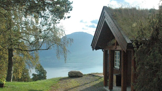 En rustik hytte med træfacade, veranda og udsigt til sø og fjelde i baggrunden.