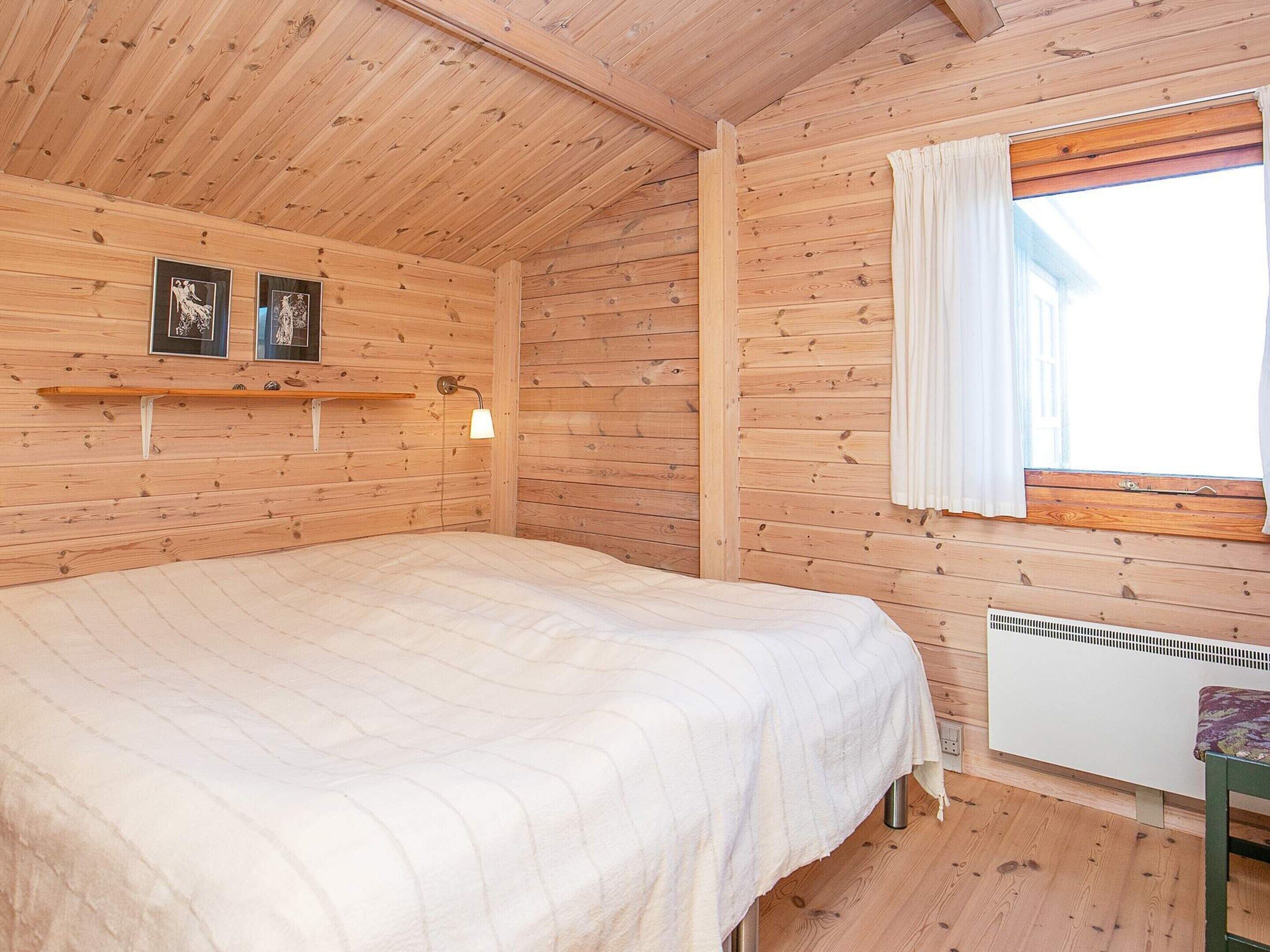 A wooden cabin room with a bed, a window with curtains, and a wall-mounted heater.
