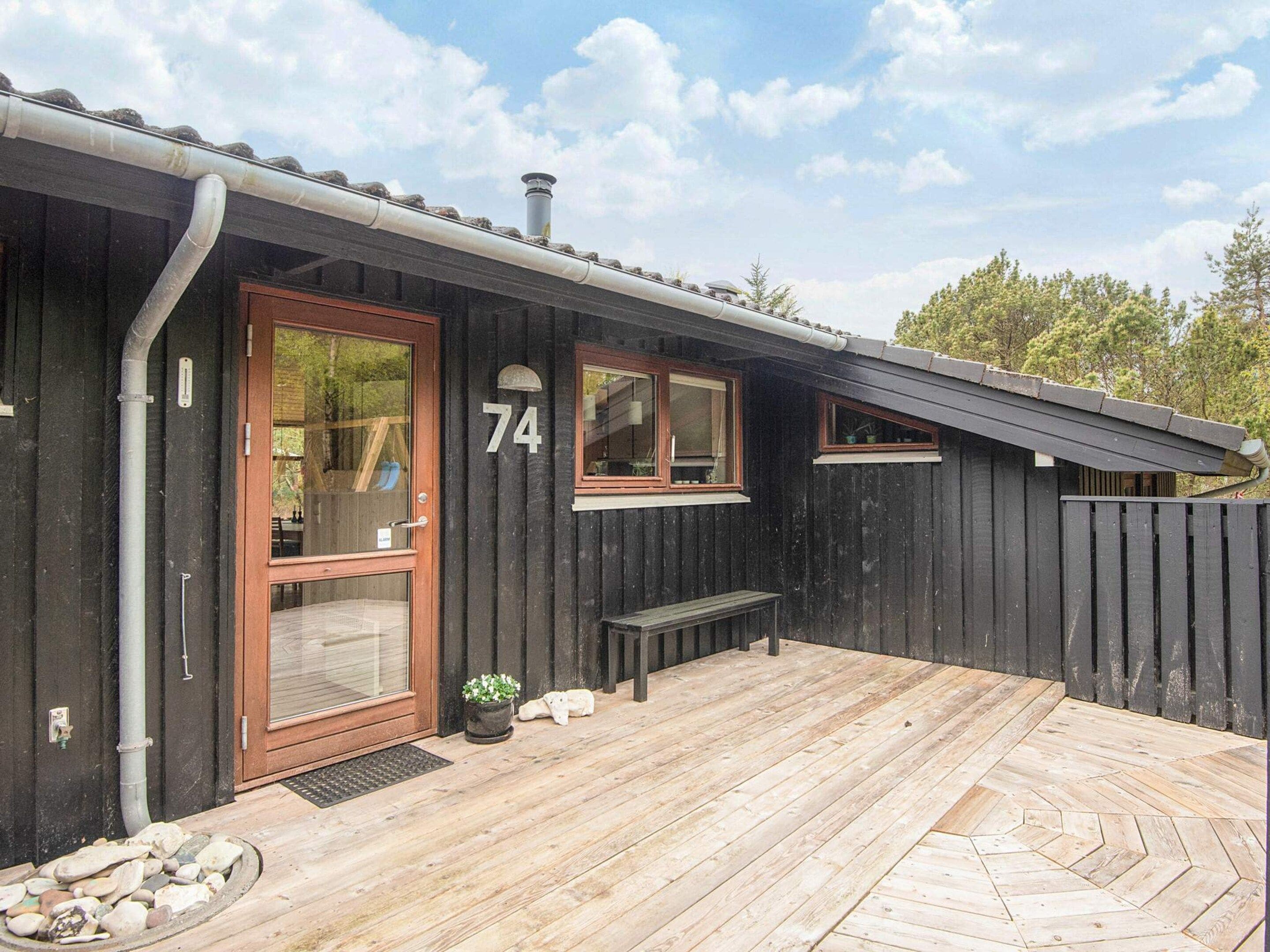 A wooden cabin with a number 74, a front door, windows, a bench, and a wooden deck.