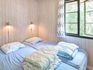 Una habitación con dos camas, cada una con almohada y manta. Hay luces fijadas a la pared y una ventana con cortinas.