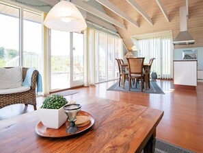 Un vaste espace de vie comprenant une table à manger en bois, un canapé en osier et de grandes fenêtres donnant sur l’extérieur.