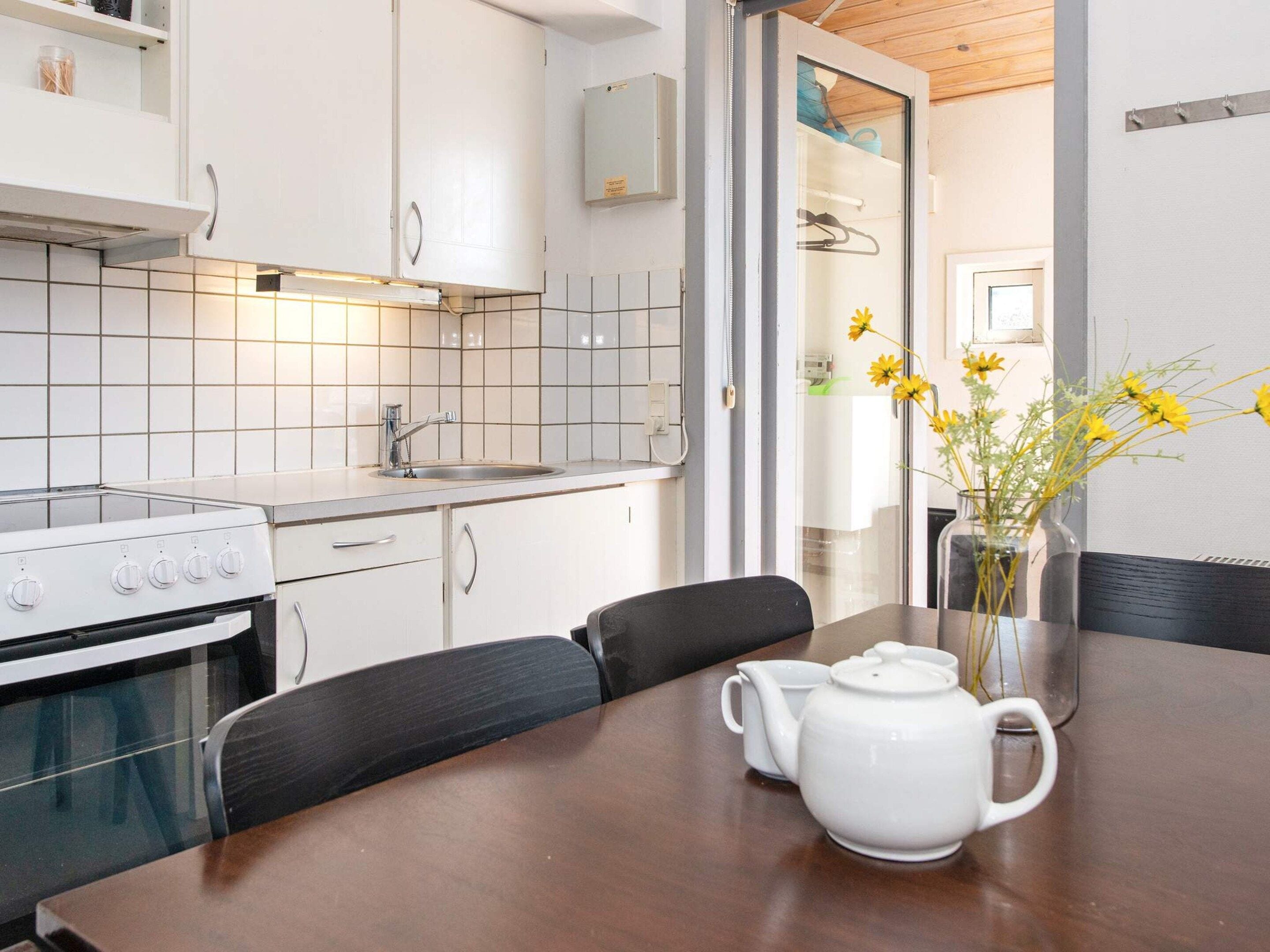 A modern kitchen with white cabinets, a stove, and a sink. There is a wooden table with a white teapot and a vase of yellow flowers.
