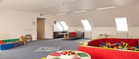 Een ruime speelkamer met een hoge plafond, dakramen en diverse speeltoestellen en tafels.