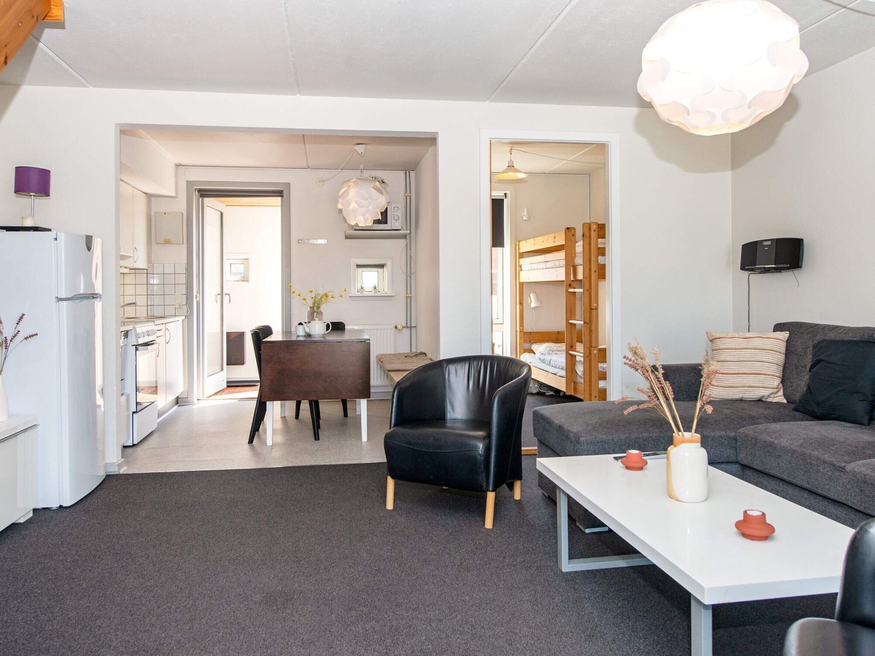 A modern living room with a sofa, a dining table, and a black leather chair. There is a kitchen area with a refrigerator and a stove.