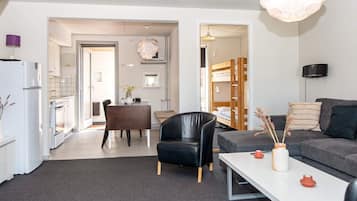 A modern living room with a sofa, a dining table, and a black leather chair. There is a kitchen area with a refrigerator and a stove.