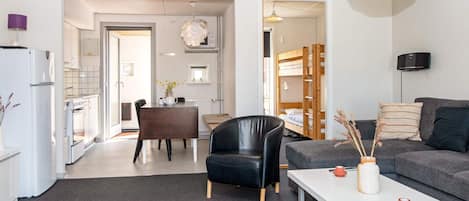 A modern living room with a sofa, a dining table, and a black leather chair. There is a kitchen area with a refrigerator and a stove.