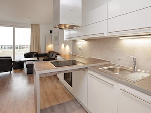 A modern kitchen with integrated appliances, a dining area with a sofa and table, and a view through large windows. - Holiday Home in Wendtorf (Wendtorf)