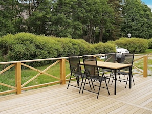 A wooden deck with a table and chairs, surrounded by greenery and a white vehicle in the background.