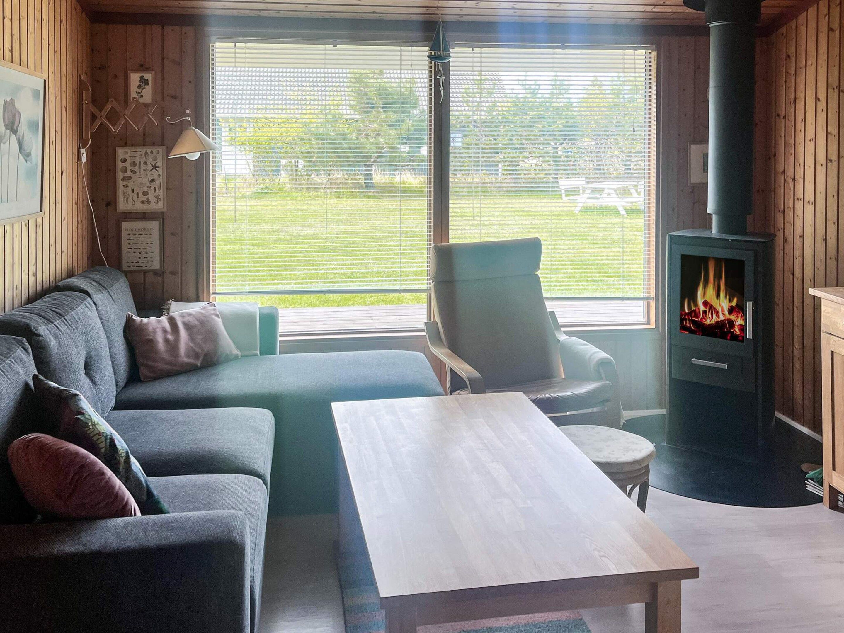 Un salón acogedor con sofá, sillón y una mesita. Se ve una chimenea y ventanas con persianas.
