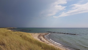 Ein Strand mit einer grasbewachsenen Vordergrund, einem steinernen Wellenbrecher und einem ruhigen Meer unter einem teils bewölkten Himmel.
