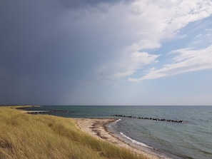 A beach with a grassy foreground, a rocky breakwater, and a calm sea under a partly cloudy sky. - 4 Person Holiday Home in Rodby (Rodby)