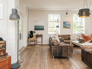 A cozy living room with a wooden floor, a leather sofa, a wooden side table, and a large window with a view.