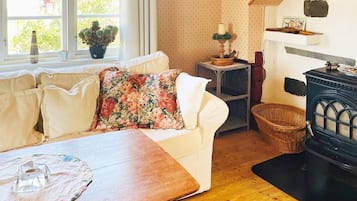 Una habitación acogedora con una mesa de madera, un sofá blanco con cojines estampados, una ventana con plantas y una estufa negra.