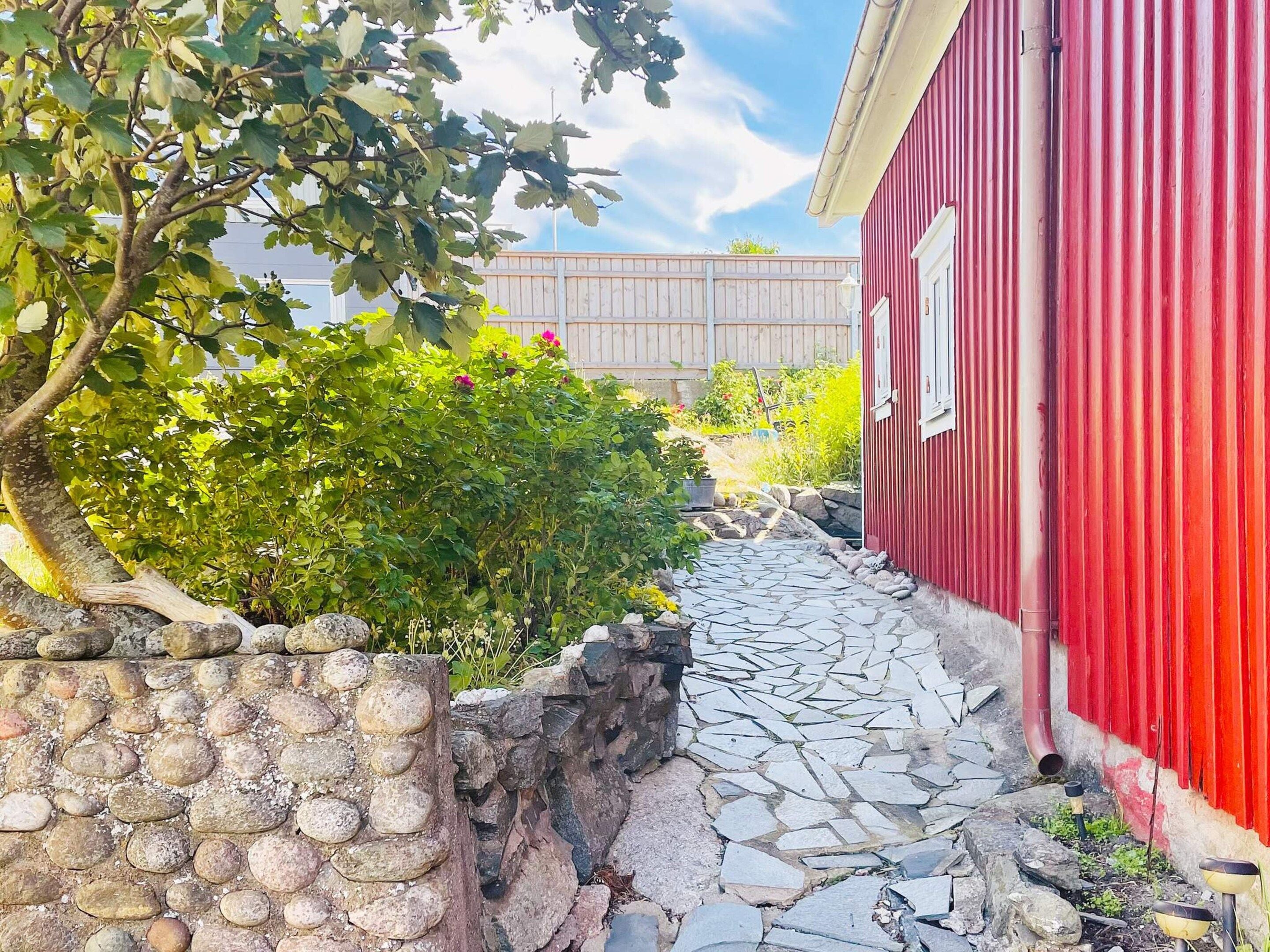 A stone pathway leading to a red building with a window, surrounded by greenery and a wooden fence.