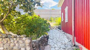 Un sendero de piedra conduce a un edificio rojo con una ventana, rodeado de vegetación y una cerca de madera.
