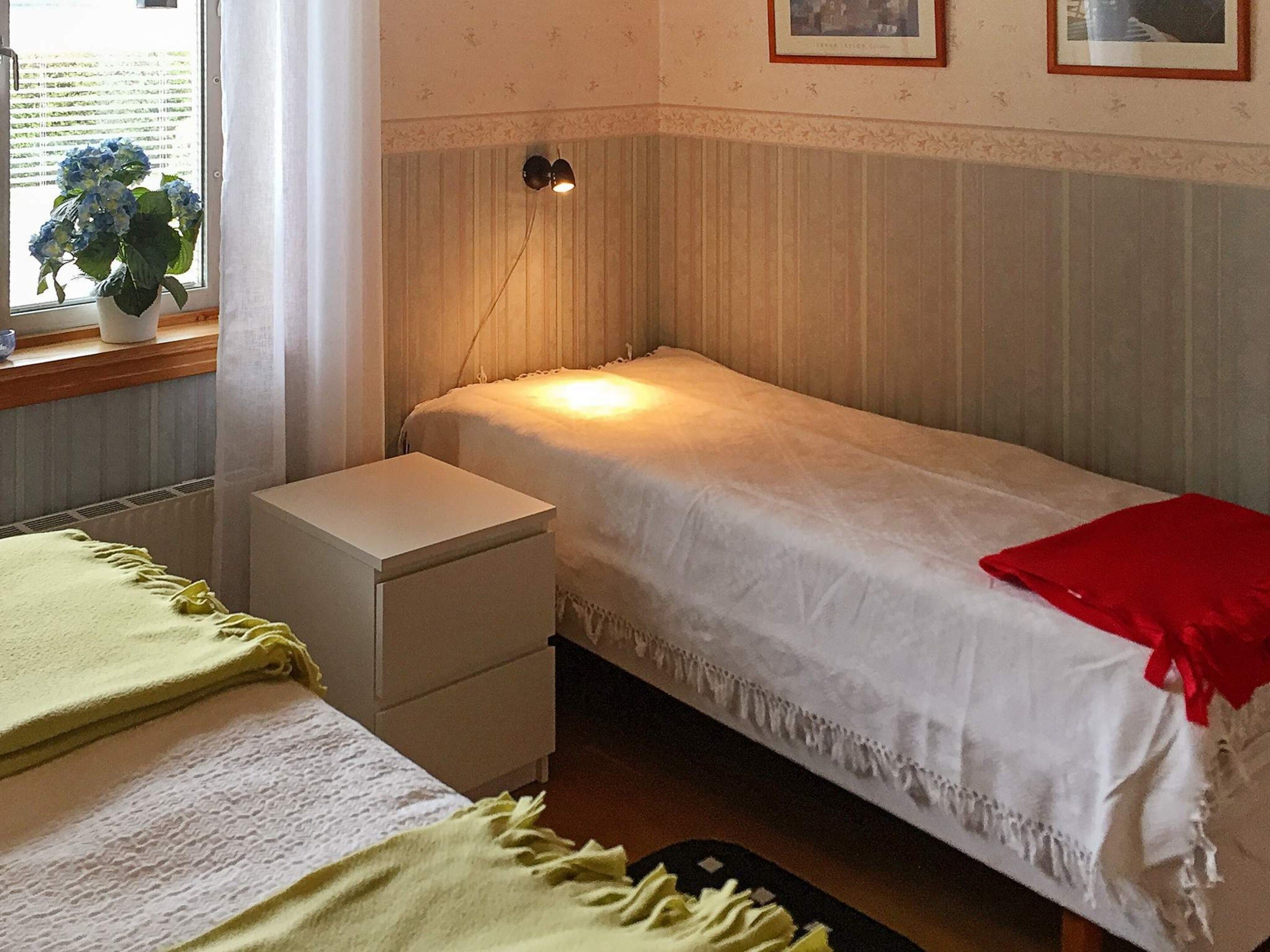 Una cama individual con una colcha roja, una mesita de noche, una lámpara y una ventana con cortinas.