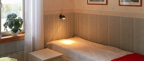 Ein Einzelbett mit roter Decke, ein Nachttisch, eine Lampe und ein Fenster mit Vorhängen.