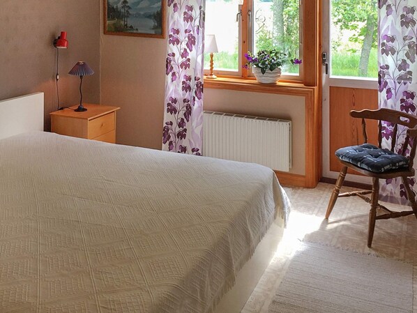 Une chambre à coucher comprenant un lit, une table de chevet, une chaise, une fenêtre avec des rideaux et un radiateur.