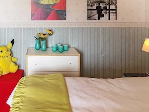 Une chambre d’hôtel avec un lit, une table de chevet agrémentée d’objets décoratifs, un jouet en peluche à l’effigie de Pikachu et un tableau encadré accroché au mur.