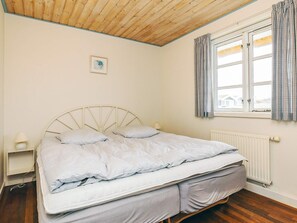 Une chambre à coucher comprenant un lit, une table de chevet, une lampe, une fenêtre avec des rideaux et un radiateur.