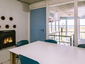 A room with a fireplace, a white table, blue chairs, and a view through a door. - 4 Star Holiday Home in Hirtshals (Hirtshals)
