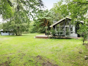 A rustic cabin with a wooden deck and a glass-enclosed porch, surrounded by lush greenery and trees.