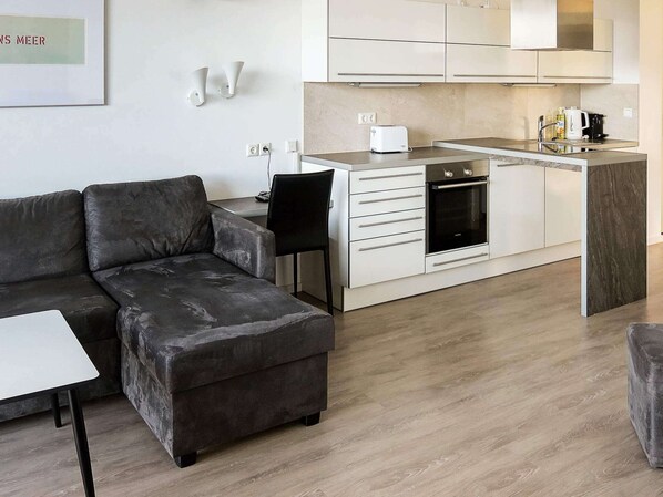A modern living room with a grey sectional sofa, a small white table, and a kitchen area with white cabinets, a built-in oven, and a toaster.