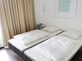 Una cama doble con ropa de cama blanca y cabecera, una mesita de noche con lámpara, y un aplique de pared.
