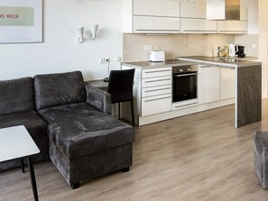 Una sala de estar moderna con un sofá seccional gris, una mesita blanca pequeña y una cocina con horno empotrado y tostadora.