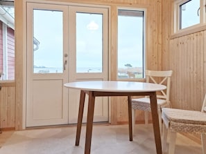 A room with a wooden interior, a round table, and two chairs. There are two doors with windows, one of which has a view of a body of water and a tree.