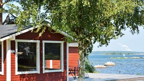 Eine rote Holzhütte mit einem Steg, der in einen See ragt, umgeben von viel Grün.