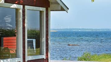 A red wooden cabin with a large window overlooking a body of water and wind turbines in the distance.